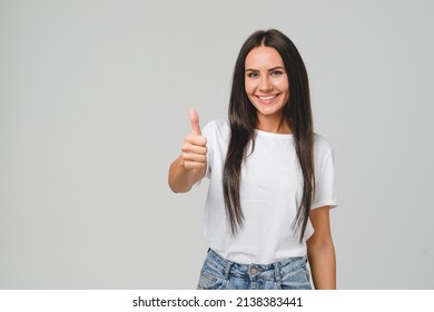 Positive caucasian young girl woman in white T-shirt showing thumb up looking at camera, checking for good quality recommendation isolated in grey background. Well done! Great job! - Shutterstock ID 2138383441