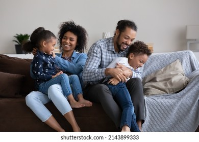 Positive cheerful Black mom and dad cuddling, tickling two children on sofa giggling, laughing, enjoying family fun, home activities, leisure, being parents. African couple playing with preschool kids
