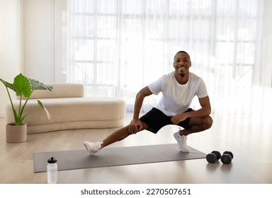 Positive cheerful handsome millennial african american guy in sportswear doing squats on fitness mat, black man enjoying workout at home, smiling at camera, copy space - Powered by Shutterstock