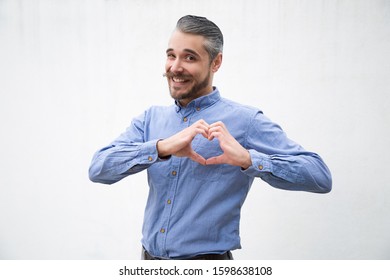 Positive cheerful man making heart with hands. Grey haired young man in blue casual shirt posing isolated over white background. Love gesture concept - Shutterstock ID 1598638108