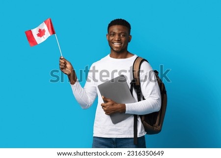 Positive cheerful millennial african american guy student with backpack holding laptop and Canadian flag over blue studio background. Education abroad, student visa to Canada