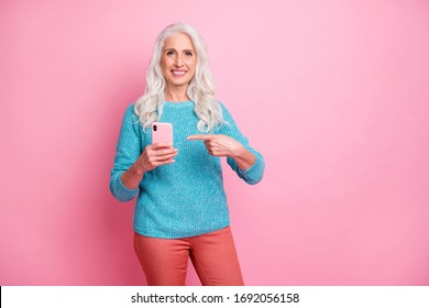 Positive cheerful old woman point index finger smartphone advise choose decide buy new modern technology device wear casual pullover red pants trousers isolated pastel color background