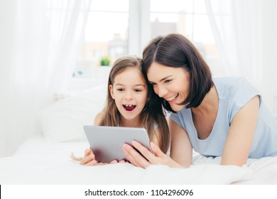 Positive Cheerful Small Female Kid And Her Young Mother Watch Interesting Cartoon On Tablet, Connected To Wireless Internet, Pose Against Bedroom Interior, Have Happy Expressions. Family Concept