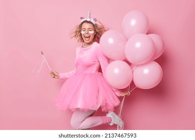 Positive cheerful young woman exclaims from joy keeps mouth widely opened poses with bunch of inflated balloons dances and has fun moves energetic against pink background. Party time concept