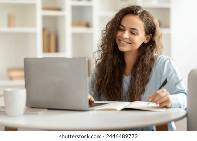 Positive cheerful young woman student using laptop and taking notes, attending webinar, have online training at home interior - Powered by Shutterstock