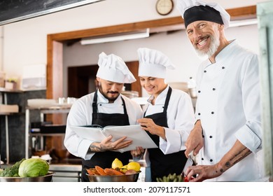 Chef positivo con cuchillo cerca de colegas multiétnicos en delantales leyendo libros de cocina y verduras en la cocina