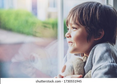 Positive Child Boy Sitting At The Window Writing On Glass And Looking Out With Smiling Face, School Kid Self Isolation Relaxing At Home During Qualrantine Covid 19, Home Schooling , Distance Education