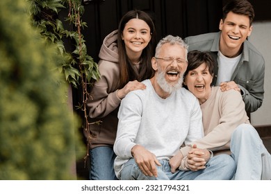 Positive children looking at camera while hugging middle aged parents holding hands and sitting on porch of house during parents day celebrating , parent-child relationship concept
