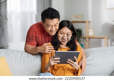 Positive chinese spouses using digital tablet at home, having video call with friends, happy asian man hugging his pretty wife, looking at pad screen and smiling, copy space. Technologies and leisure