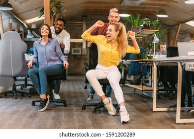 Positive Colleagues Having Fun In Office, Enjoying Office Chair Race And Competing