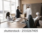 Positive confident Arab and European businessmen shaking hands at team meeting, standing at large table, talking, smiling, laughing, discussing contract, deal, partnership