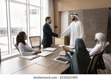 Positive confident Arab and European businessmen shaking hands at team meeting, standing at large table, talking, smiling, laughing, discussing contract, deal, partnership