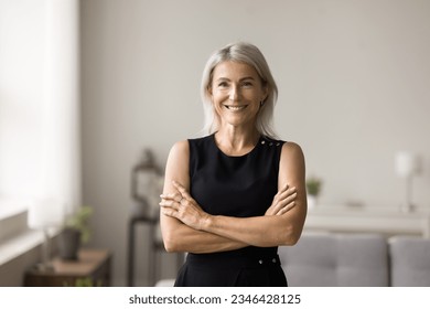 Positive confident blonde mature woman wearing black sleeveless cloth, standing at home interior, looking, smiling at camera with crossed arms, laughing. Successful senior entrepreneur portrait - Powered by Shutterstock