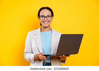 Positive Confident Clever Lovely Latino Or Brazilian Young Brunette Woman, Business Lady, With Glasses, Hold In Hands Open Laptop, Stand On Isolated Orange Background, Looks At Camera, Smiles Friendly