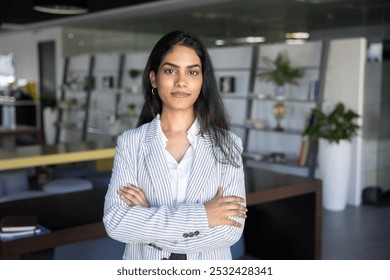 Positive confident Indian entrepreneur woman looking at camera with hands folded, standing in co-working space. Beautiful young female professional, businesswoman, company leader business portrait