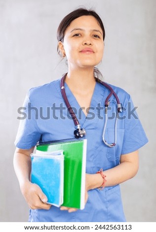 Positive confident young woman, professional doctor, in blue uniform and with phonendoscope around neck holding medical records. Studio photoshoot on gray background