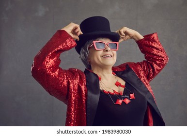 Positive Confident Youthful Looking Grandma Having Fun At A Party. Happy Beautiful Senior Woman In Black Top Hat, Cool Stylish Glasses, Jewelry And Red Sequin Concert Outfit Posing On Gray Background