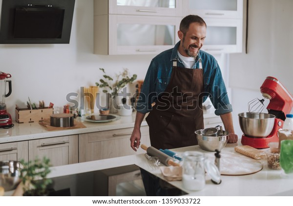 Positive cook. Emotional adult man standing in\
the kitchen and wearing an apron while looking at the electric\
stand mixer and\
laughing