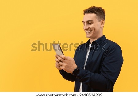 Positive cool european guy using smartphone isolated on yellow studio background, texting friends, using newest mobile app, scrolling, copy space for advertisement