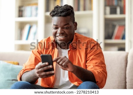 Positive cool stylish overweight young black guy with dreadlocks sitting on couch in living room, using smartphone at home, chatting with friends, websurfing, using nice app, copy space