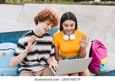 Positive couple boy girl teenagers students high school pupils classmates bloggers sitting in skate park ramp using holding laptop streaming live online have video zoom call with friends waving hands - Powered by Shutterstock