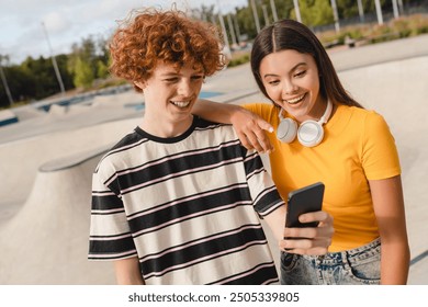 Positive couple of college students high school pupils teenagers boy girl holding smart phone watching video using mobile app play game posting chatting texting spend time in skate park - Powered by Shutterstock