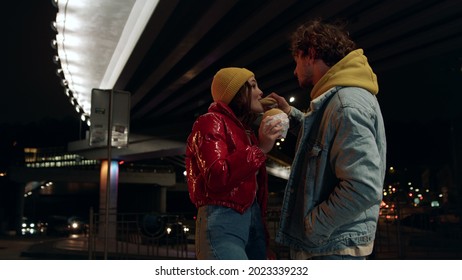 Positive Couple Eating Burgers Under Bridge At Night. Happy Man Feeding Woman During Romantic Date On Urban Background. Cheerful Young Pair Having Snacks Food Outdoor. 