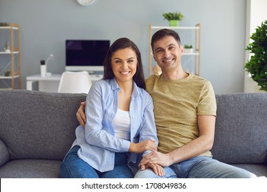 A Positive Couple Embracing Looking At The Camera While Sitting On A Sofa In The Interior Room Of The House.