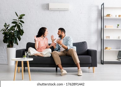 Positive couple smiling at each other while sitting on sofa under air conditioner at home