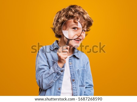 Positive curious schoolboy in casual wear looking at camera through magnifier while standing against bright orange background