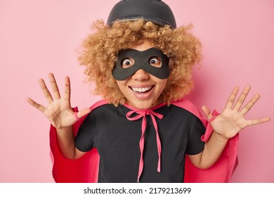 Positive Curly Female Heroine Dressed In Masquerade Costume Keeps Palms Raised Entertains Someone At Party Looks Gladfully Pretends To Have Extraordinary Ability Isolated Over Pink Background