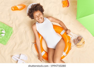 Positive Curly Haired Afro American Woman Has Fun Holds Net Bag Of Fruits Poses In Lifebuoy Wears Panama And White Swimsuit Spends Free Time At Sandy Beach During Summer Holidays. Overhead Shot