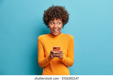 Positive curly haired ethnic woman uses mobile phone checks messages and reads news holds modern cellular in hands looks with curious happy expression on right isolated over blue background.