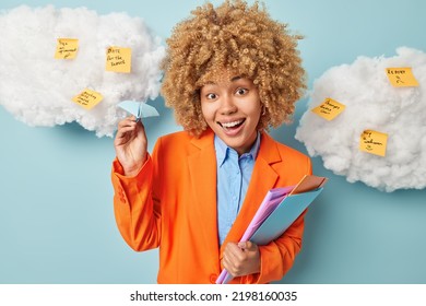 Positive Curly Haired Woman Dressed In Orange Formal Jacket Uses Stickers To Write List To Do Holds Paper Handmade Aeroplane And Folders Poses Against Blue Background With White Clouds Around