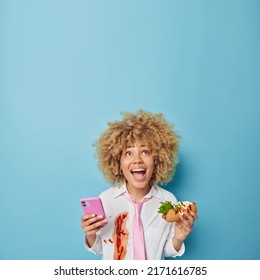 Positive Curly Haired Woman Holds Mobile Phone And Hot Dog Eats Unhealthy Harmful Food Focused Above With Glad Surprised Expression Dressed Formally Isolated Over Blue Background With Empty Space