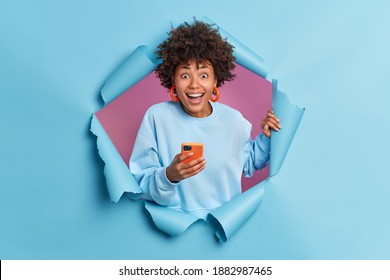 Positive curly haired woman smiles broadly uses smartphone reads funny news on website reacts on something awesome and pleasant dressed in casual jumper poses in ripped hole. Staying always in touch