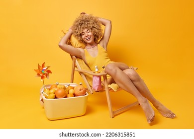 Positive Curly Haired Young Woman Dressed In Swim Suit Poses On Comfortable Deck Chair Laughs Happily Keeps Eyes Closed Enjoys Summer Vacations At Beach Uses Portabe Fridge Full Of Citrus Fruits