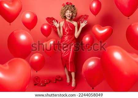 Positive curly woman keeps hands raised smiles broadly wears festive dress and shoes prepare for celebrating St Valentines Day stands in full length against red background with heart shaped balloons