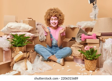 Positive Curly Woman Sits Crossed Legs On Floor Applies Beauty Mask On Face Prepares For Party Moves To New Apartment Surrounded By Cardboard Boxes Full Of Household Items Rents House For Living