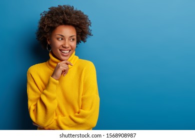 Positive curly young woman dressed in yellow comfortable sweater, holds chin, looks aside with dreamy expression, has interesting idea in mind, isolated over blue background. Ethnicity concept
