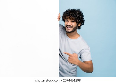 Positive curly-haired Indian or Arabian guy, in casual t-shirt, peeking out from behind advertisement white board, demonstrating blank copy space for your text or design, on isolated blue background