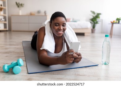 Positive Curvy Black Woman Using Smartphone During Her Break From Home Training, Browsing Internet, Looking Up Workout Video Online. Overweight Afro Lady Checking Cellphone After Exercising