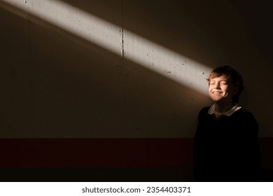 Positive cute preteen boy standing with closed eyes at wall in dark room catching sunbeam in face