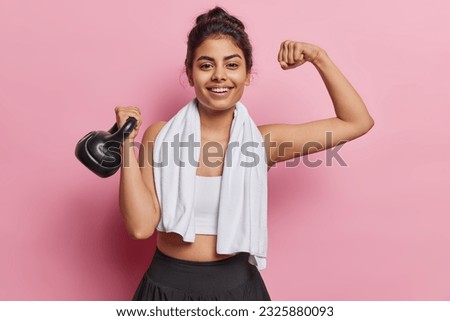 Positive dark haired sporty young woman raises arm shows muscles lifts weight stands satisfied smiles gladfully dressed in sportswear poses with towel around neck isolated over pink background