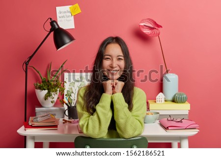 Positive dark haired woman with pleasant smile, keeps hands under chin, studies at home against desktop, prepares for upcoming exams, isolated over pink background, happy to complete homework
