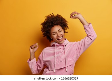 Positive dark skinned curly woman keeps hands raised in air, dances carefree, feels lively and upbeat, wears velvet hoodie, isolated over yellow studio background, throws party, enjoys freedom - Shutterstock ID 1666820329