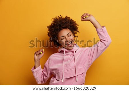Positive dark skinned curly woman keeps hands raised in air, dances carefree, feels lively and upbeat, wears velvet hoodie, isolated over yellow studio background, throws party, enjoys freedom Foto stock © 
