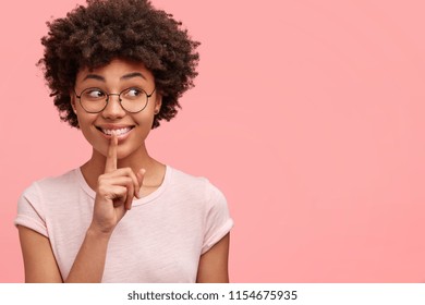Positive dark skinned female with curly bushy hair, has toothy smile, makes hush gesture, wears eyeglasses and casual clothes, poses against pink studio wall with copy space for your advertisement