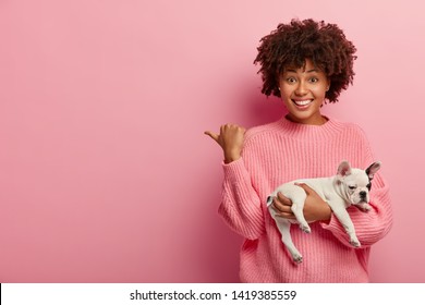 Positive Dark Skinned Lady Points Thumb Away, Wears Casual Jumper, Holds Sleeping Dog, Being Caring Hostess, Shows Direction To Animal Shop, Isolated Over Pink Background. Pedigree Puppy With Owner