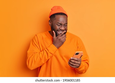 Positive dark skinned man giggles positively concentrated at smartphone screen watches funny video in internet or laughs over received message dressed in casual bright clothes isolated on orange wall